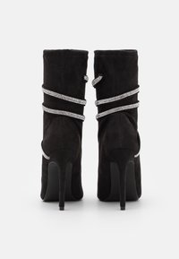 Bottines en suede noir avec un talon haut. Présente deux sangles métalliques argentées ornées de strass enroulées autour de chaque bottine.