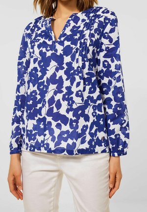 Vrouw draagt een witte blouse met lange mouwen met blauwe bloemenprint en een witte broek, getoond van nek tot heup tegen een effen achtergrond.