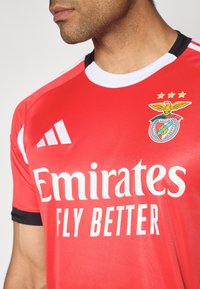 Rød trøje med sorte detaljer og hvid krave, der har mærket "Emirates Fly Better" samt et klublogo med en ørn og tre stjerner.