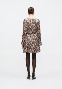 Γυναίκα με μαλλιά σε κότσο φορούσα φόρεμα με μακριά μανίκια, με σχέδιο και βολάν, μαύρο καλσόν και μαύρα παπούτσια με τακούνι, στέκεται μπροστά σε λευκό φόντο.