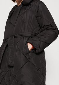 Osoba v černém prošívaném péřovém kabátu s páskem, s jednou rukou v boční kapse, pod kterou má hnědý rukáv.