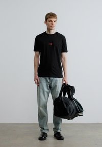 Μαύρο κοντομάνικο μπλουζάκι με μικρό ορθογώνιο γραφικό, σε συνδυασμό με ανοιχτό μπλε τζιν παντελόνι και μαύρα παπούτσια, κρατώντας μια σκούρα τσάντα duffel.