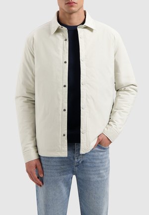 Hellbeige Button-up-Jacke mit Kragen, Kontrastnähten und vorderen Taschen, getragen über einem dunklen Marinepullover und Jeans.