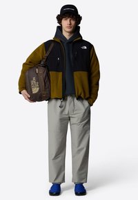 Giacca in pile con pannelli neri e oliva, felpa grigia con cappuccio, pantaloni leggeri grigi e scarpe slip-on blu. Borsa a tracolla marrone con logo.