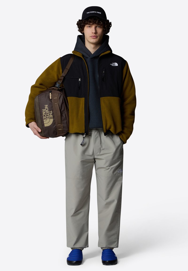 Giacca in pile con pannelli neri e oliva, felpa grigia con cappuccio, pantaloni leggeri grigi e scarpe slip-on blu. Borsa a tracolla marrone con logo.