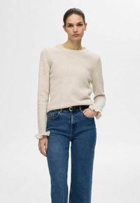 Pull en tricot beige avec un col rond et des poignets à volants, associé à un jean droit taille haute bleu. Texture douce, coupe décontractée.