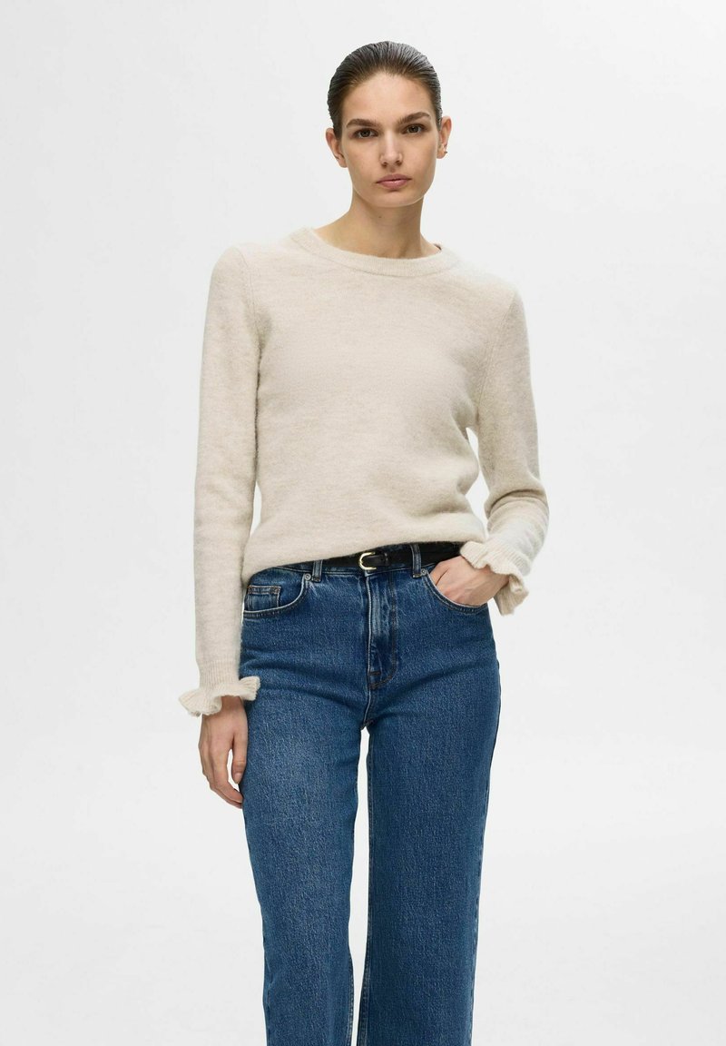 Pull en tricot beige avec un col rond et des poignets à volants, associé à un jean droit taille haute bleu. Texture douce, coupe décontractée.