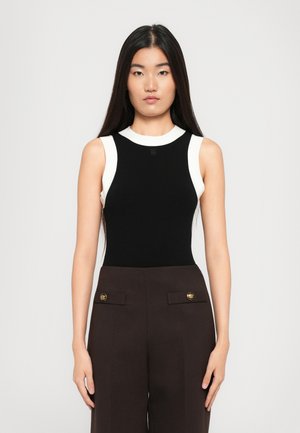 Mujer con largo cabello negro lacio, vistiendo una camiseta sin mangas negra con ribetes blancos y pantalones de tiro alto marrón oscuro con botones dorados.