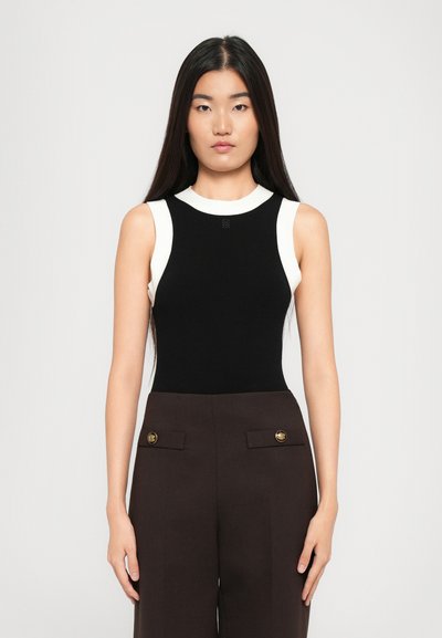Frau mit langen, glatten schwarzen Haaren, die ein ärmelloses schwarzes Top mit weißer Borte und hoch taillierte, dunkelbraune Hose mit goldenen Knöpfen trägt.