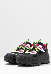 Scarpe sportive in bianco e nero con tomaia in rete, lacci verdi e fodera rosa. Suola spessa in gomma con motivo di trazione testurizzato.