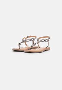 Sandales plates argentées métalliques avec des brides tressées, fermeture à la cheville et semelle tan, présentant un design minimaliste et un bout ouvert.