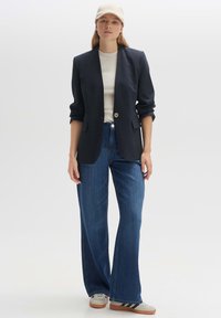 Blazer marine à bouton unique, associé à un haut crème et un jean bleu à jambes larges. Les accessoires comprennent une casquette beige et des baskets.