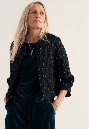 Femme aux longs cheveux blonds portant une tenue en velours vert foncé et une veste noire à motifs, regardant sur le côté les mains dans les poches.