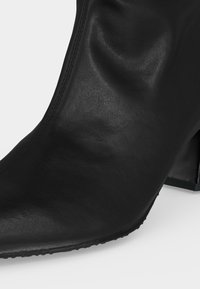 Gros plan d'une botte d'cheville en cuir noir avec un bout pointu et un talon bloc sur fond blanc.
