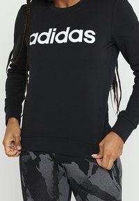 Personne portant un sweat à capuche noir Adidas avec un logo blanc et des leggings à motifs noir et gris, tenant l'ourlet du sweat.