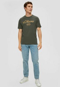 Olive green t-shirt with short sleeves, featuring brown text "s.Oliver Elevated Essentials" on the front; paired with light blue jeans and white sneakers.
