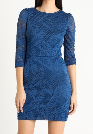 Niebieska koronowana sukienka z wzorami paproci, dopasowaną talią, przezroczystymi rękawami o długości do łokcia oraz okrągłym dekoltem na stojącej kobiecie.