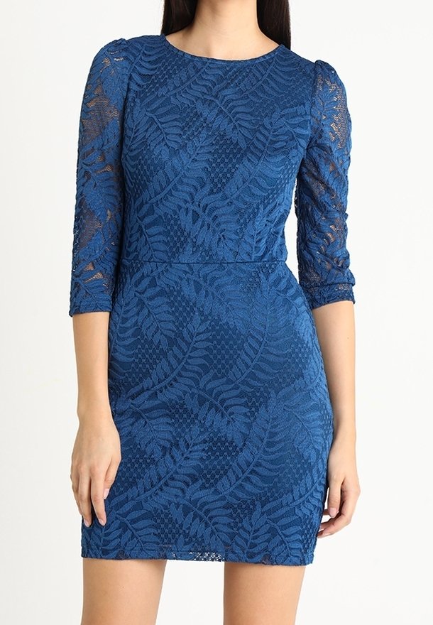 Robe en dentelle bleue avec motifs de fougères, taille cintrée, manches trois-quarts transparentes et encolure ronde portée par une femme debout.