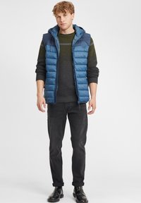 Vadderad blå väst med huva, har en texturerad mörkgrön och grå tröja under, tillsammans med svarta jeans och svarta stövlar.