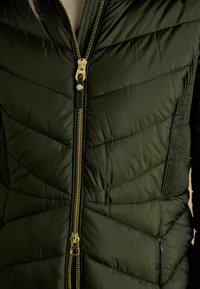 Tmavozelená páperová bunda s chevronovým dizajnom, zlatým zipsom a štruktúrovanými bočnými panelmi. Zips prebieha vertikálne po celej dĺžke.