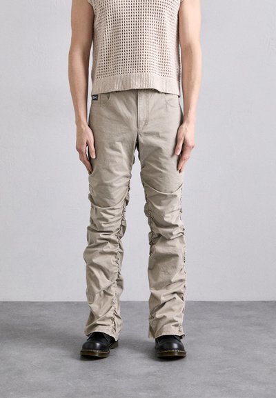 Pantalons cargo beiges à poches multiples avec texture froncée et détails élastiques, portés avec des bottes noires. Portés par une personne devant un fond neutre.