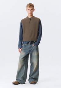 Jovem vestido com um sweater castanho sem mangas sobre uma camisa de manga longa azul, jeans largos desbotados, sapatos castanhos e cinto preto, em pé contra um fundo branco.