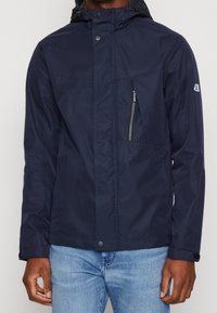 Navy blue waterproof jacket with a hood, featuring a zippered chest pocket, snap buttons, and textured fabric. Standard fit with long sleeves.