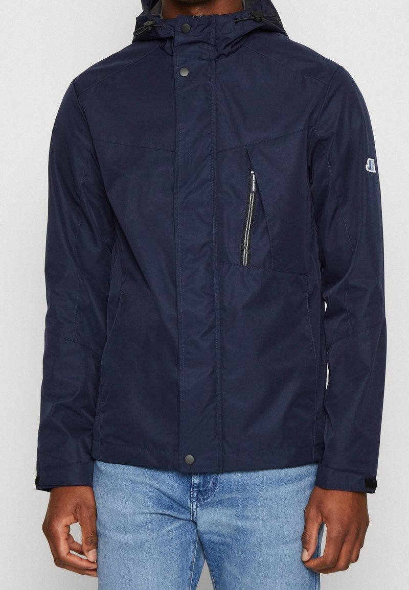 Navy blue waterproof jacket with a hood, featuring a zippered chest pocket, snap buttons, and textured fabric. Standard fit with long sleeves.