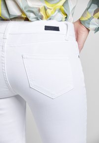 Nahaufnahme einer Person in weißen, eng anliegenden Jeans und einem bunten, gemusterten Hemd. Die hintere Tasche und die Hand, die auf der Hüfte ruht, sind zu sehen.
