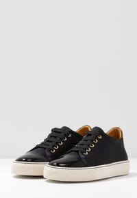 Sneakers neri a bassa altezza con punta in vernice liscia, tomaia in tela, occhielli dorati, lacci in tessuto e suola in gomma testurizzata di colore bianco sporco.