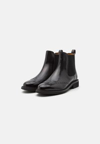 Bottes Chelsea en cuir noir avec perforations brogues sur les embouts et panneaux latéraux élastiques, présentées sur un fond clair uni.