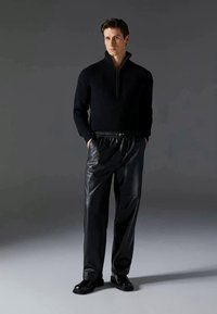 Suéter preto de malha canelada com fecho, combinado com calças pregueadas de couro pretas e sapatos pretos, apresentando um ajuste descontraído e um design estruturado.