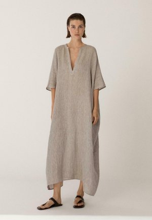 Frau trägt ein lockeres, strukturiertes, hellbeiges knöchellanges Kleid mit tiefem V-Ausschnitt und halblangen Ärmeln, kombiniert mit schwarzen Sandalen.