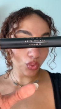 Personne aux cheveux bouclés tient un bâton de maquillage noir étiqueté « GLDR Shadow Stick Cocoa Drip » près de son visage, les lèvres pincées.