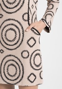 Vestido beige con destacados patrones circulares y en forma de diamante en negro. Cuenta con mangas largas y bolsillos laterales, con un acabado de tela texturizada.