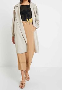 Beige trenssitakki musta-keltaisella kuviolla olevan paidan päällä, yhdistettynä ruskeisiin leveälahkeisiin housuihin ja ruskeisiin nauhallisiin sandaaleihin.