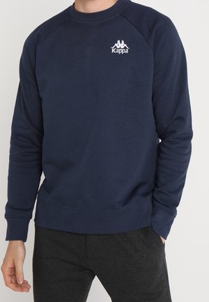 Mężczyzna w granatowej bluzie Kappa z długimi rękawami i ciemnoszarych spodniach, stojący z jedną ręką w kieszeni.