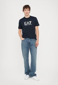 T-shirt in cotone blu navy con logo bianco "EA7 EMPORIO ARMANI", abbinata a jeans dritti blu chiaro e sneakers bianche.