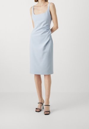 Frau trägt ein ärmelloses, hellblaues Midikleid mit quadratischem Ausschnitt und schwarze Sandalen mit hohen Absätzen, steht vor einem schlichten Hintergrund.