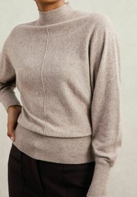 Beige pletený svetr s vysokým límcem a žebrovanými manžetami. Má šev probíhající vpředu a přiléhající pas. Měkká textura.