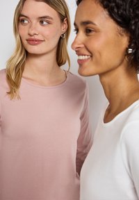 Twee vrouwen in nauwsluitende lange mouwen tops. De ene in zachtroze met een ronde hals, de andere in wit met een wijde hals. Beide hebben een gladde stof.