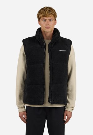 Gilet in pile nero con colletto alto, chiusura a zip e tasche a toppa. Indossato sopra una camicia a maniche lunghe beige con texture.
