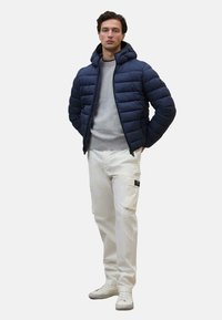 Giacca a vento blu navy con cappuccio, maglione grigio, pantaloni cargo bianchi e sneakers bianche. La giacca ha un design trapuntato e chiusura con zip.