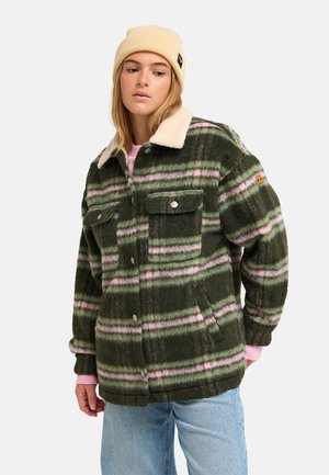 Junge Frau trägt eine grün-rosa gestreifte, flauschige Jacke, beige Mütze und hellblaue Jeans und schaut leicht zur Seite.