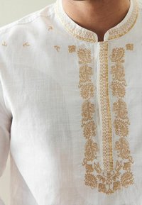 Uomo che indossa una camicia tradizionale bianca con ricami dorati a motivo paisley e floreale lungo il colletto e la patta anteriore.