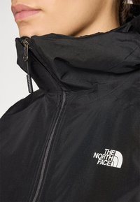 Black softshell jacket with a high collar, front zipper, and white logo on the chest. Features smooth texture and lightweight design.