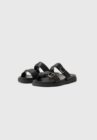 Black sandals with two wide straps and a gold circular accent. Flat sole with textured edge, made from smooth synthetic material.