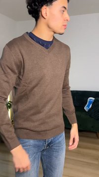 Brūns džemperis ar V veida kaklu, ribotiem manžetiem un apakšmalu. Valkāts virs tumšas krekla, kopā ar zilām džinsa biksēm. Fonā redzama zaļa dīvāns.