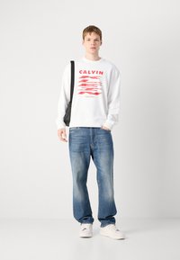 Felpa bianca con testo grafico rosso, jeans blu a vestibilità rilassata e sneakers bianche. Il modello porta anche una borsa a tracolla nera, in piedi davanti a uno sfondo neutro.