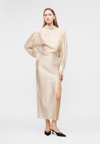 Γυναίκα με μακρυμάνικο μπλουζάκι από μπεζ σατέν και φούστα midi σετ με σχίσιμο στο μπούτι, φοράει μαύρες μυτερές γόβες, ποζάρει σε λευκό φόντο.